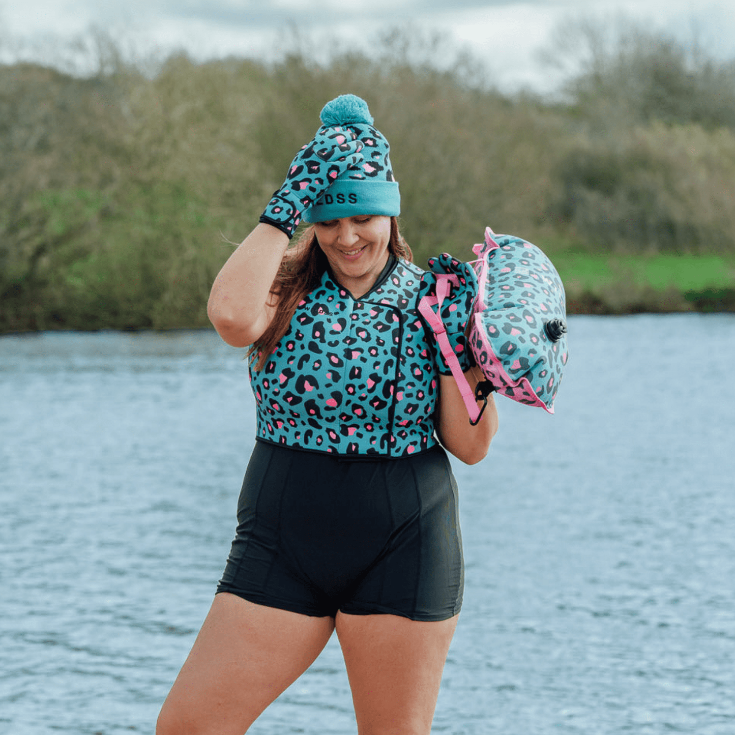 Person wearing a leopard print neoprene body warmer and woolly hat by a body of water