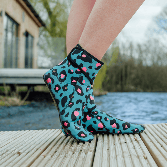Teal neoprene socks with pink and black leopard print worn by a person on a wooden dock by a body of water.