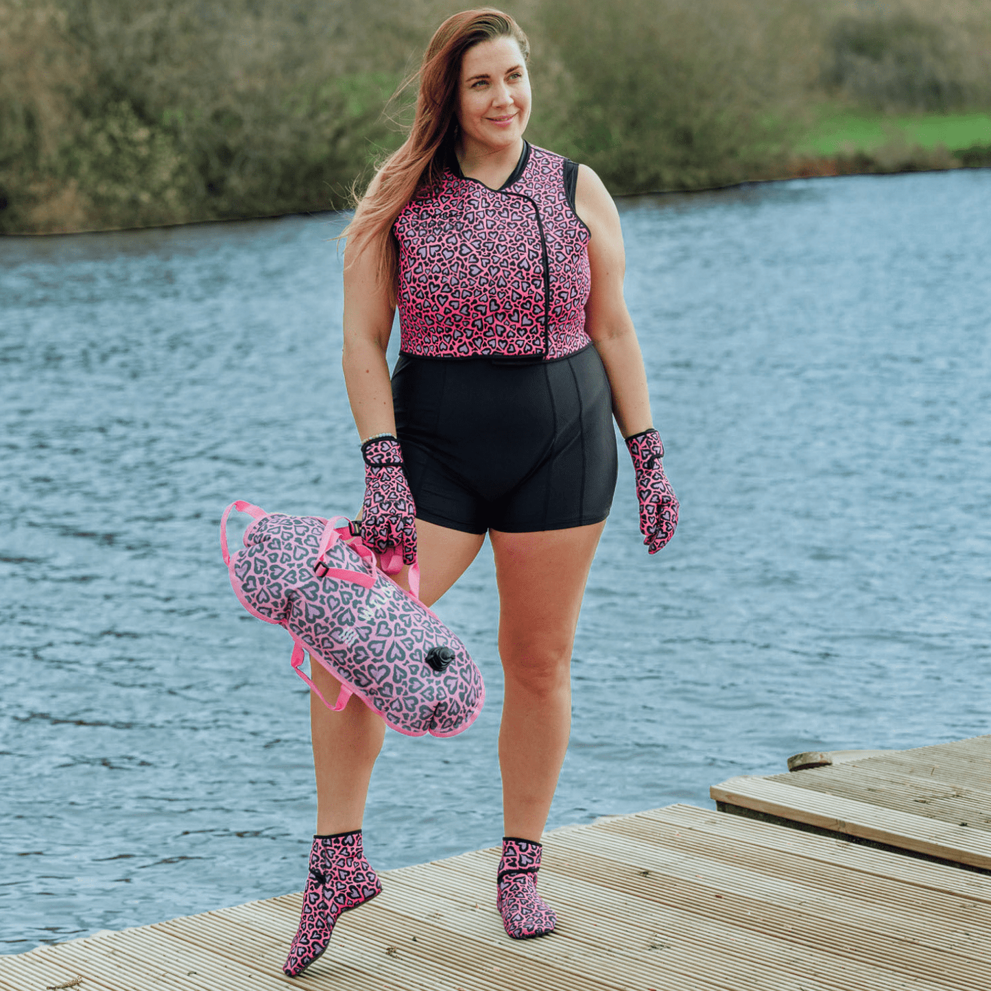 Woman stood by a lake on a dock wearing a black swimsuit and pink leopard print neoprene swimming gear