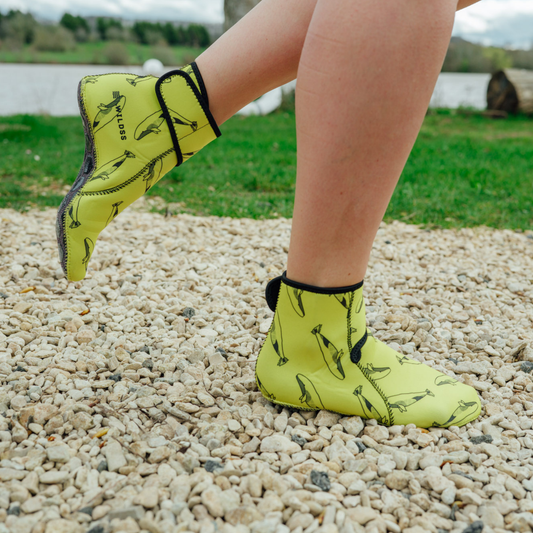 Person wearing bright yellow neoprene booties with black patterns on a gravel surface.