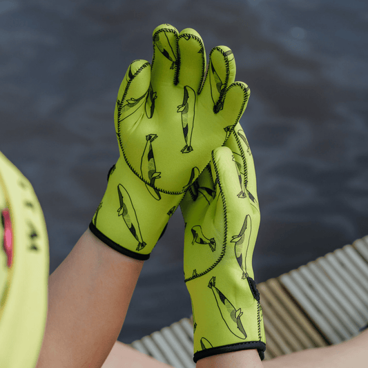 Person wearing bright yellow neoprene gloves with black whale patterns, sitting by a body of water.