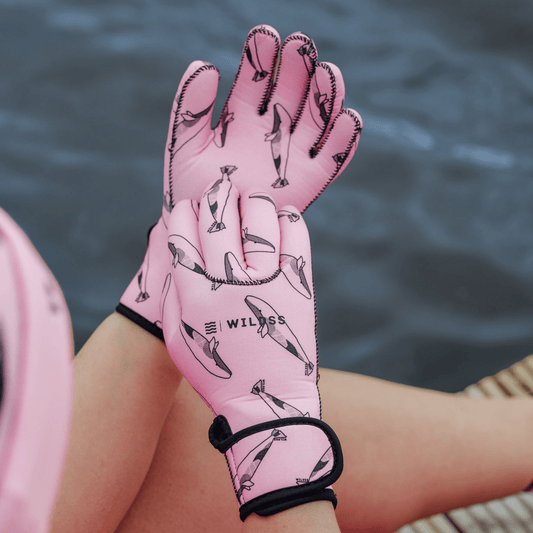Person wearing bright pink neoprene gloves with black whale patterns, sitting by a body of water.