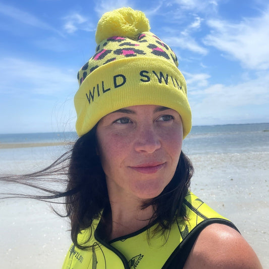 Person wearing a yellow leopard print beanie hat with 'Wild Swim' text, smiling with eyes closed outdoors