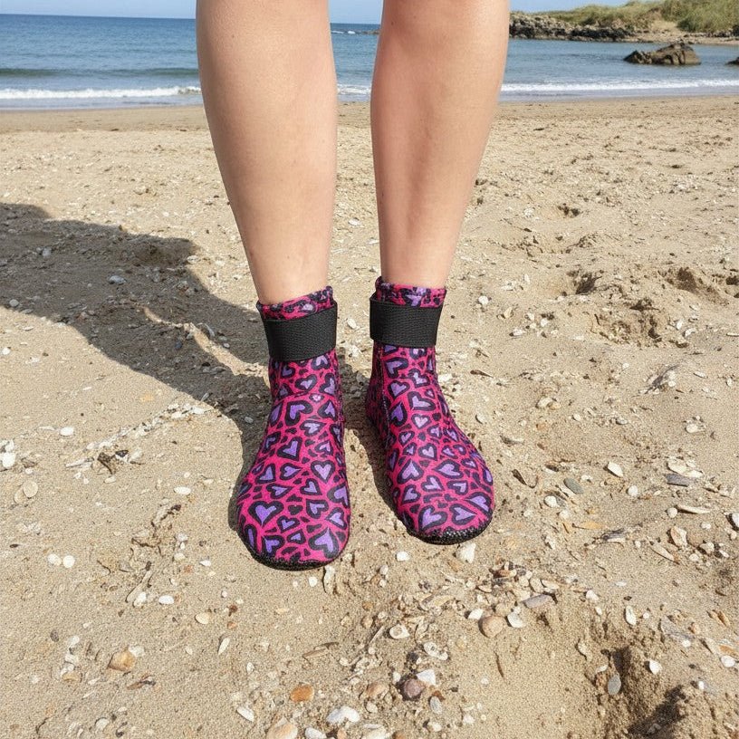 Person wearing pink and purple heart-patterned neoprene swimming socks, standing on a sandy beach with the sea in the background
