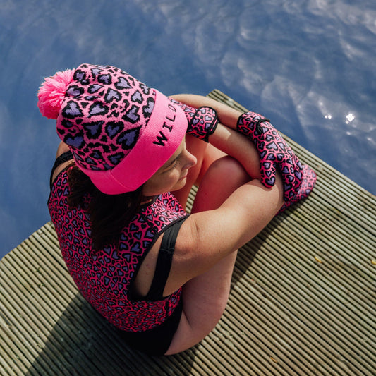 Person wearing pink and black leopard print hat and neoprene swimming gear sitting on a dock by water.
