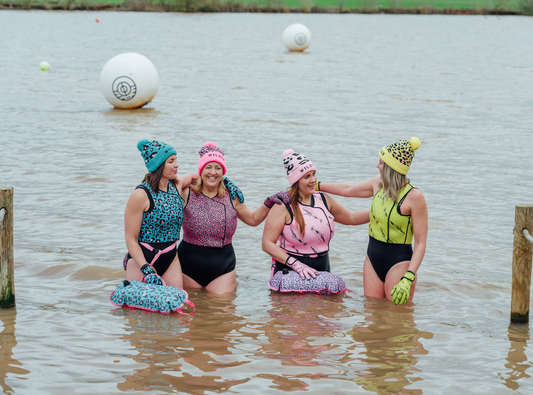 Four women stand together in a lake, smiling and chatting in colourful patterned swimwear, hats and gloves, with swim floats around them. White buoys float behind them and the far bank of the lake is visible in the background.