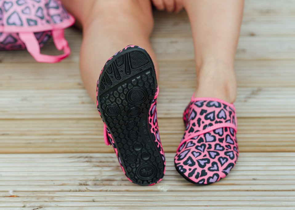 Close-up of a person sitting on a wooden dock wearing bright pink and purple heart-patterned water shoes, with a matching patterned swimsuit and swim bag beside them. One shoe is lifted to show its textured sole.