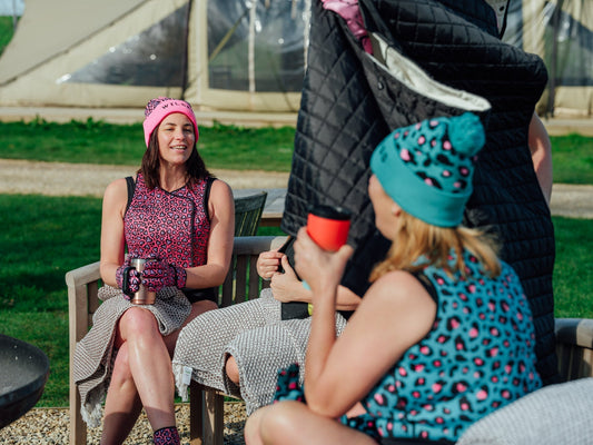 Three women in colourful patterned swimwear and beanies sit together outdoors, chatting and relaxing after a swim. One woman holds a hot drink, while towels are draped over the chairs. A grassy campsite and tents are visible in the background.