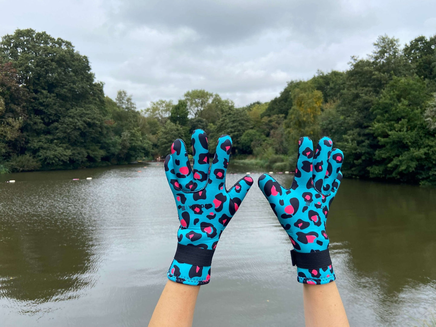Person wearing turquoise leopard print neoprene swimming gloves, hands raised by a lake
