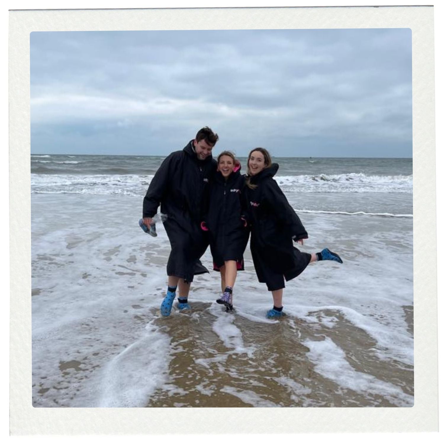 Three people in dry robes walking on a beach with waves and cloudy sky.