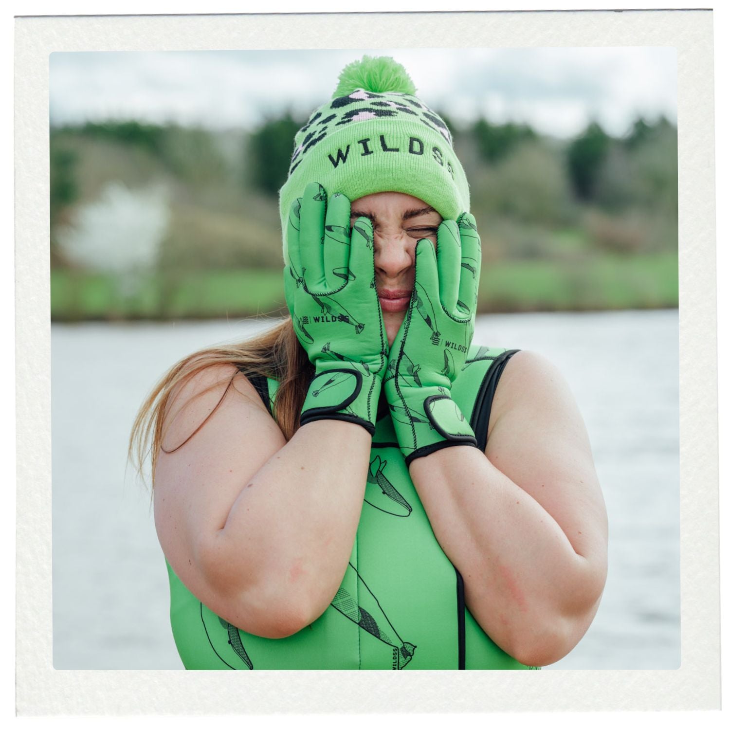 Person wearing green neoprene swimming gear with 'Wild' beanie and gloves, standing in water.