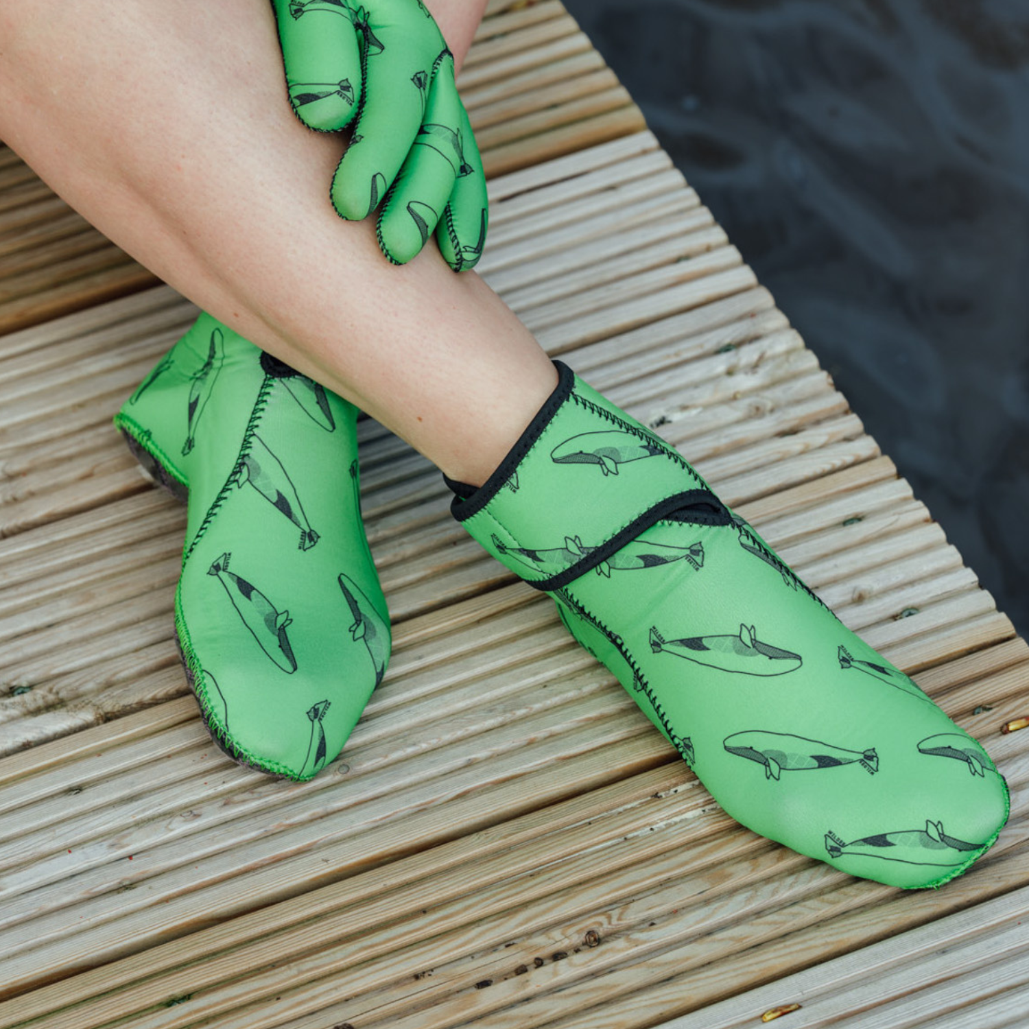 Green neoprene gloves and swimming boots with whale pattern worn by a person on a wooden surface.