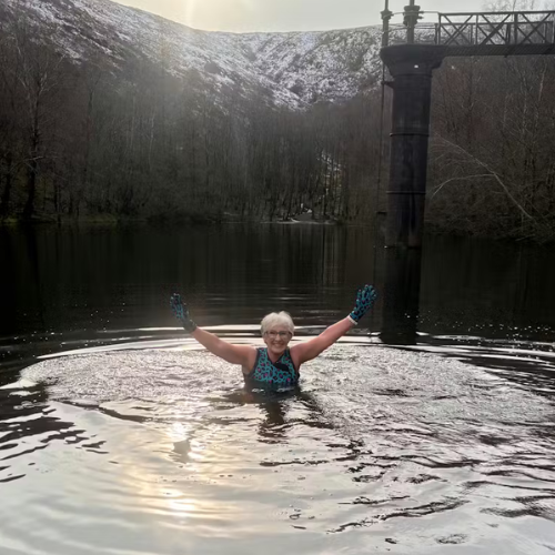 Person in a hot spring with arms raised, surrounded by snow-covered mountains.