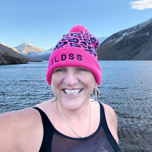 Person wearing a pink beanie with leopard print pattern standing in front of a mountain lake.