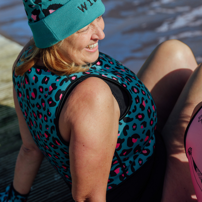 Person wearing a blue leopard print life jacket and teal beanie by a body of water.