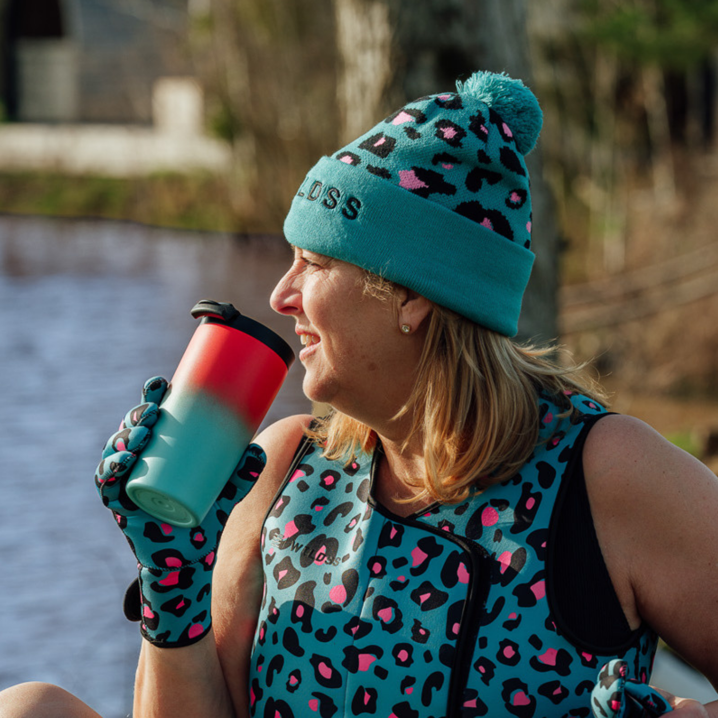 Person wearing a leopard print outfit with a teal beanie and neoprene gloves, sitting by a body of water.