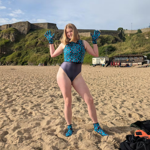 Person in a blue and black patterned swimsuit on a sandy beach with neoprene swimming gloves and matching neoprene swimming boots
