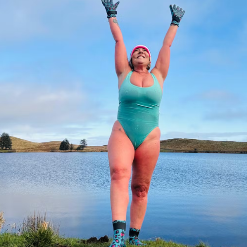 Person in a teal swimsuit standing by a lake with neoprene swimming gloves and neoprene socks with arms raised against a blue sky.