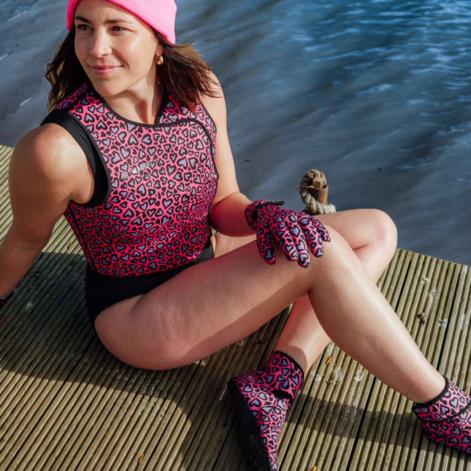 Woman sitting on a dock wearing pink and black patterned neoprene swimwear, gloves, and socks by a body of water.