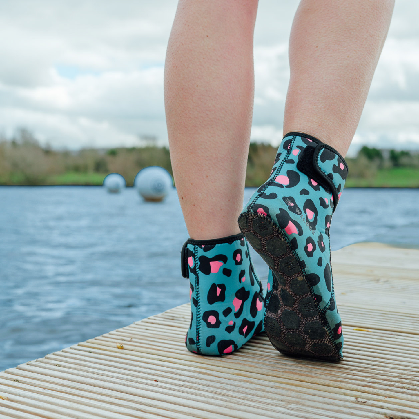 Teal neoprene beach socks with pink leopard print pattern worn by a person on a dock by water.
