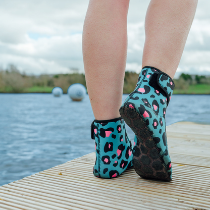 Teal neoprene beach socks with pink leopard print pattern worn by a person on a dock by water.