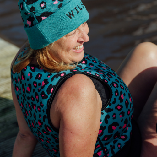 Woman sat lakeside on a dock wearing a turquoise woolly hat and neoprene body warmer