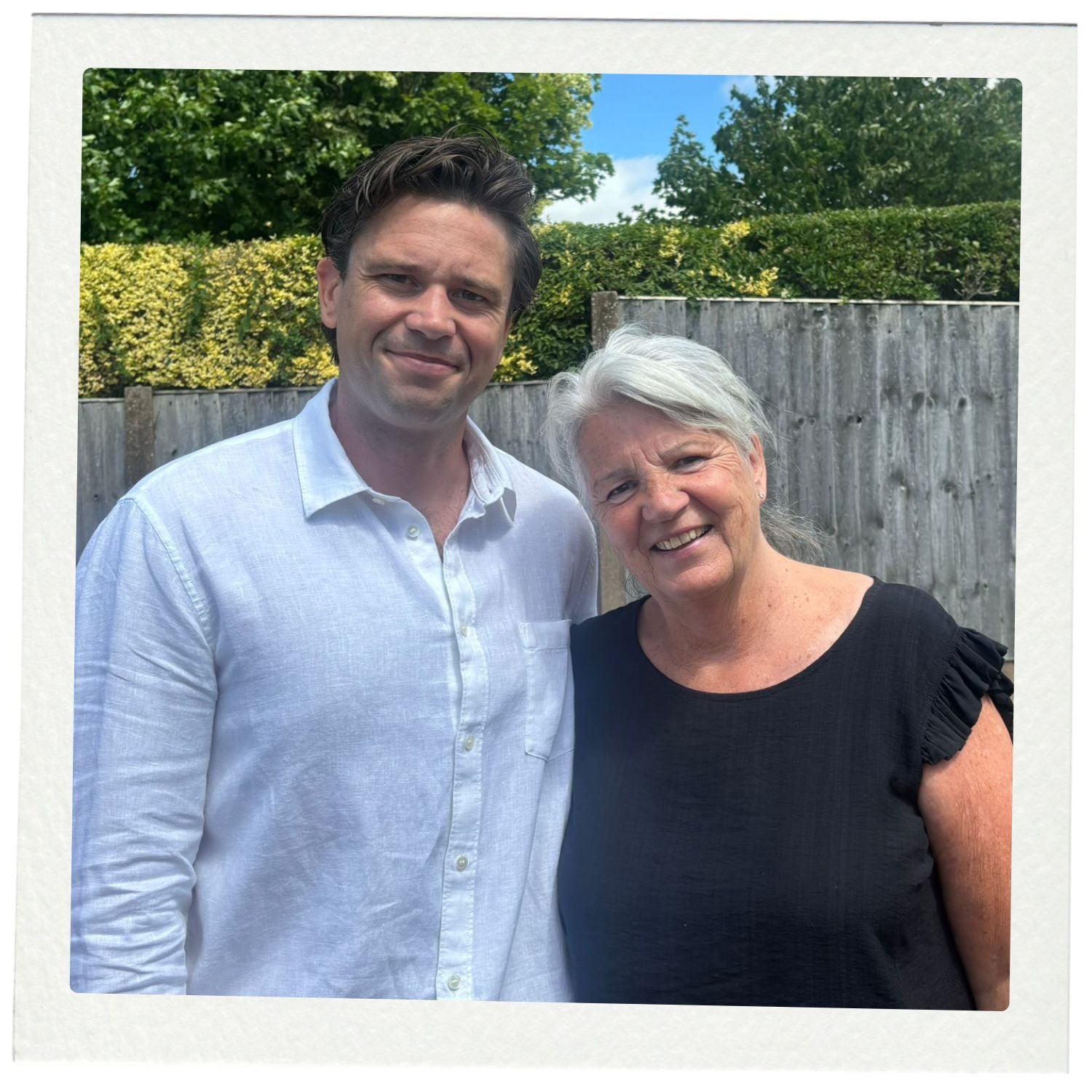 Two people standing outdoors with greenery and a wooden fence in the background