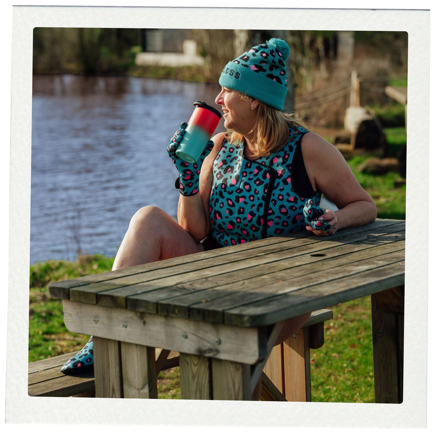 Woman sitting at a picnic table by a lake, wearing a blue patterned outfit and hat.