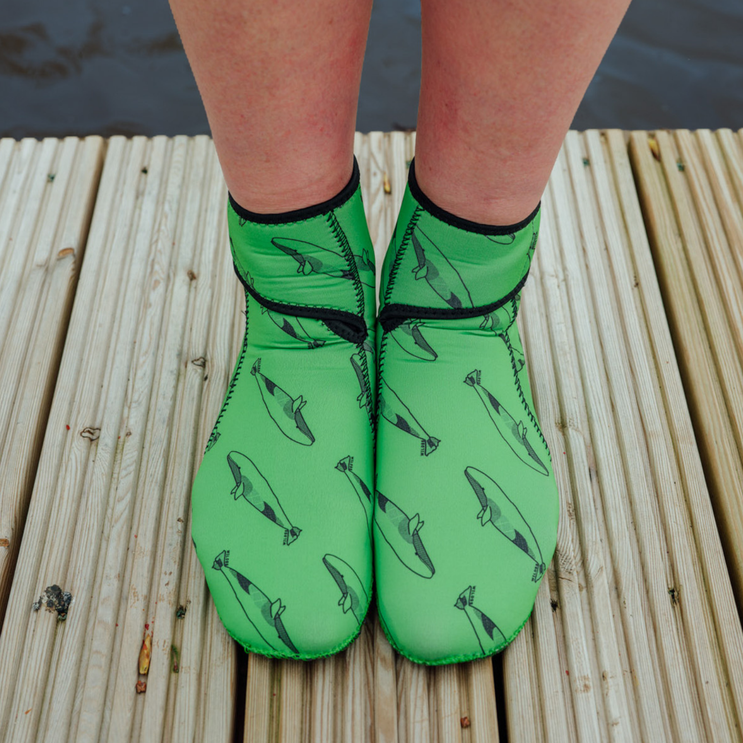 Green neoprene socks with whale pattern worn on a wooden dock.