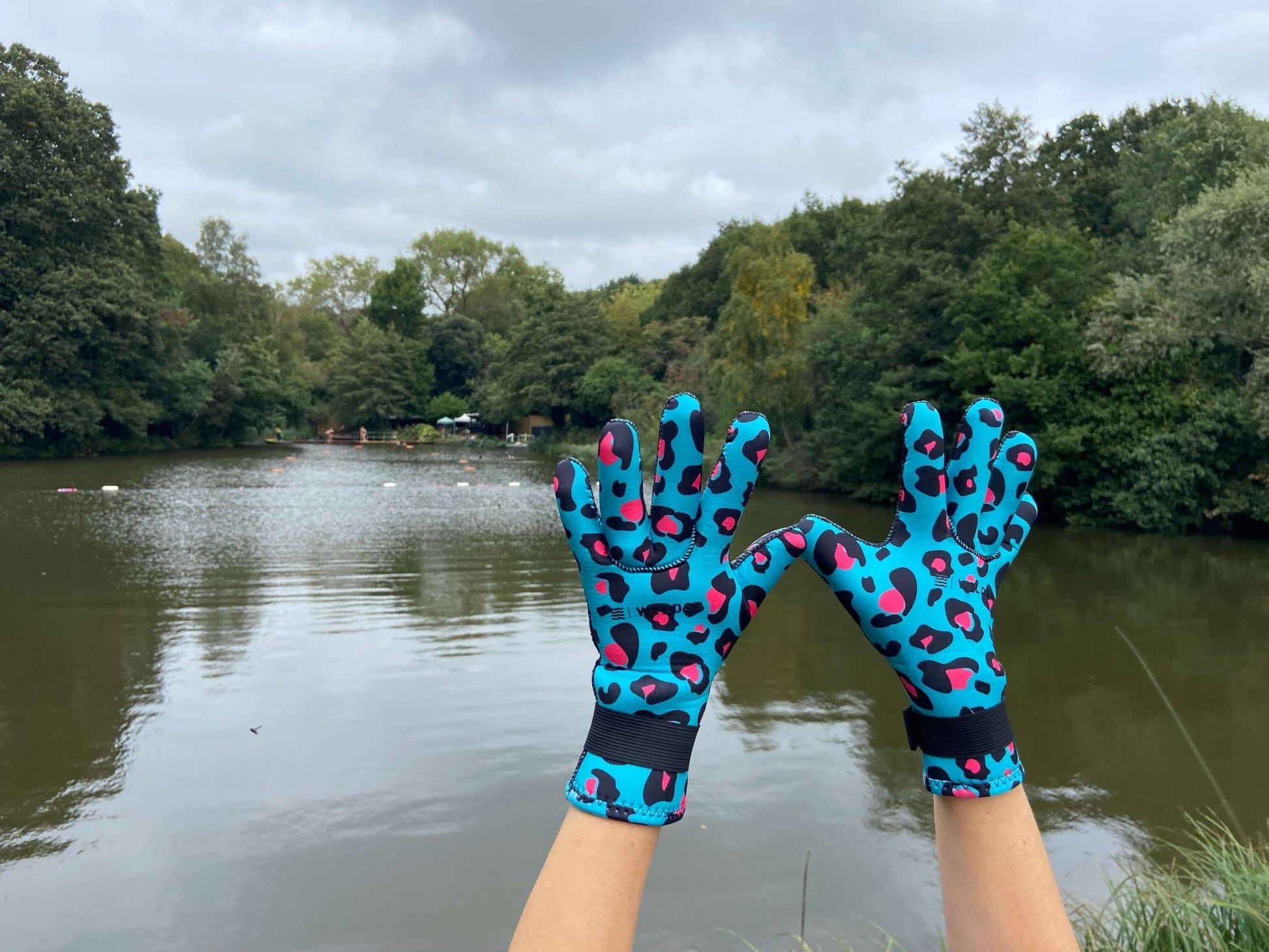 Person wearing turquoise leopard print neoprene swimming gloves, hands raised with trees in the background