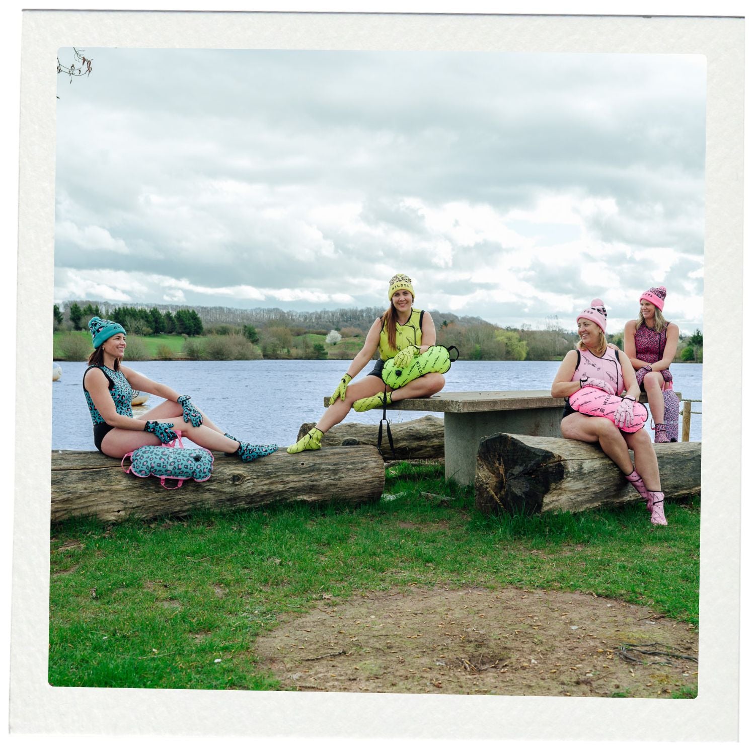 Four women in colorful outfits sitting on logs by a lake.