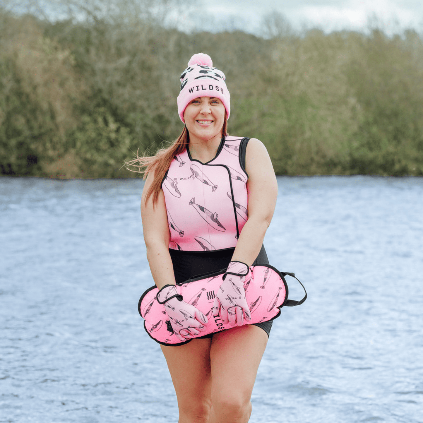 Person wearing pink hat, tow float and neoprene swimming gear by water.