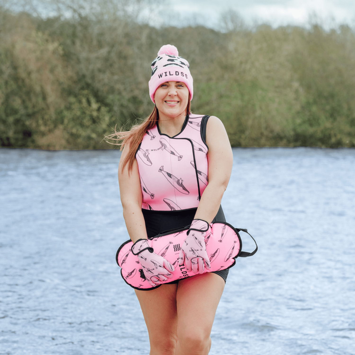 Person wearing pink hat, tow float and neoprene swimming gear by water.