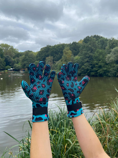 Person wearing turquoise leopard print neoprene swimming gloves, hands raised by reeds at the water's edge
