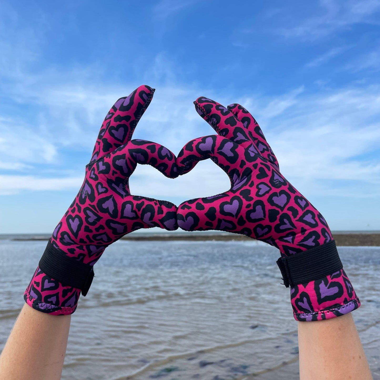 Person wearing pink and purple heart-patterned neoprene swimming gloves, hands forming a heart shape on the beach