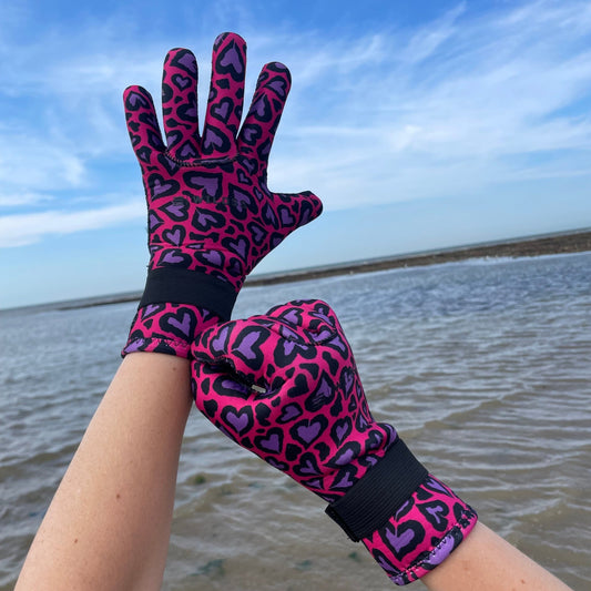 Person wearing pink and purple heart-patterned neoprene swimming gloves, one hand raised against a blue sky