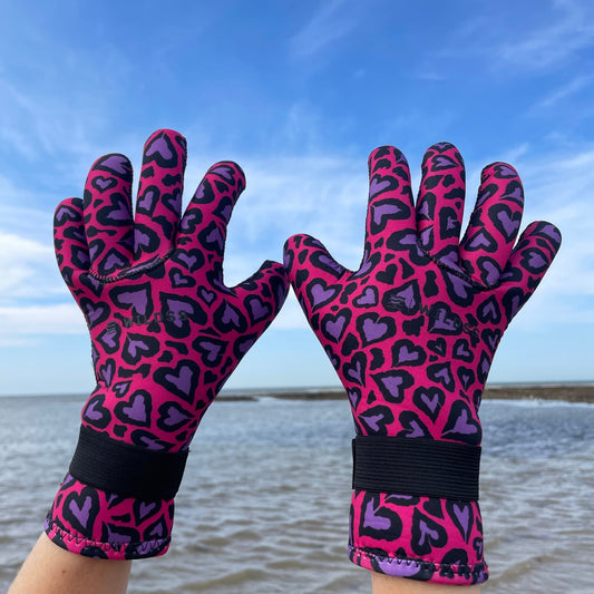 Person wearing pink and purple heart-patterned neoprene swimming gloves, hands raised with the sea in the background