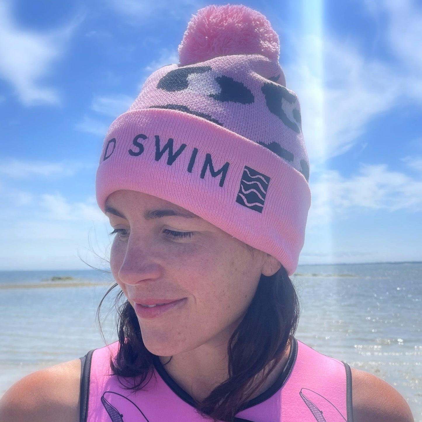 Person wearing a pink leopard print beanie hat with 'Wild Swim' text, smiling outdoors near water