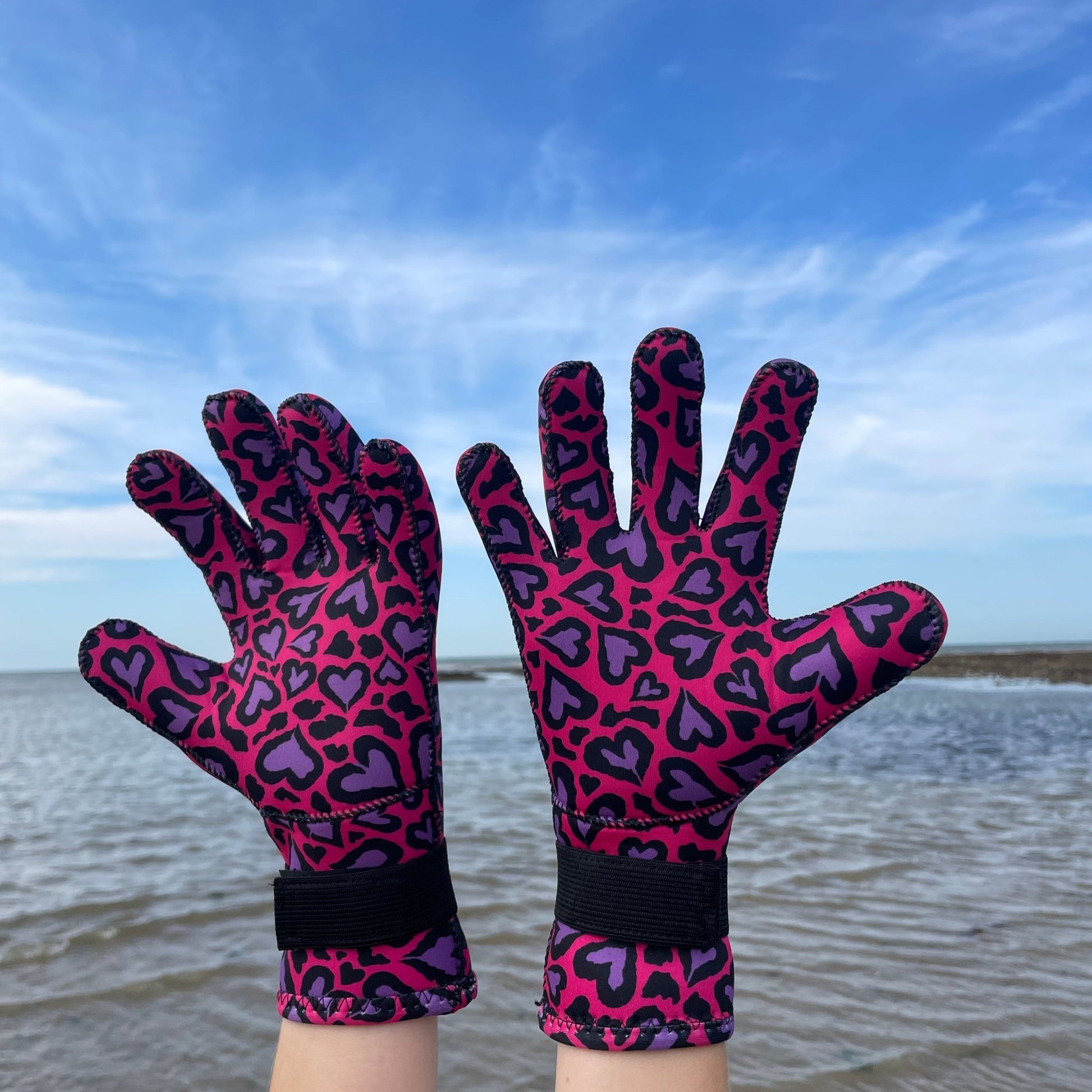 Person wearing pink and purple heart-patterned neoprene swimming gloves, hands raised by the shore