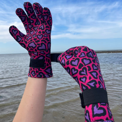 Person wearing pink and purple heart-patterned neoprene swimming gloves, hands together on the beach