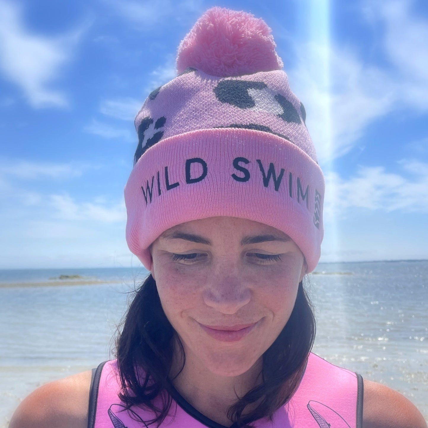 Person wearing a pink leopard print beanie hat with 'Wild Swim' text, sitting on a wooden dock by water