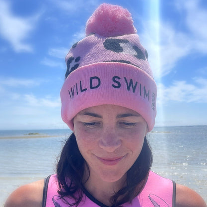 Person wearing a pink leopard print beanie hat with 'Wild Swim' text, sitting on a wooden dock by water