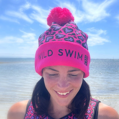Person wearing a pink leopard print beanie hat with 'Wild Swim' text, smiling and looking down outdoors by the sea
