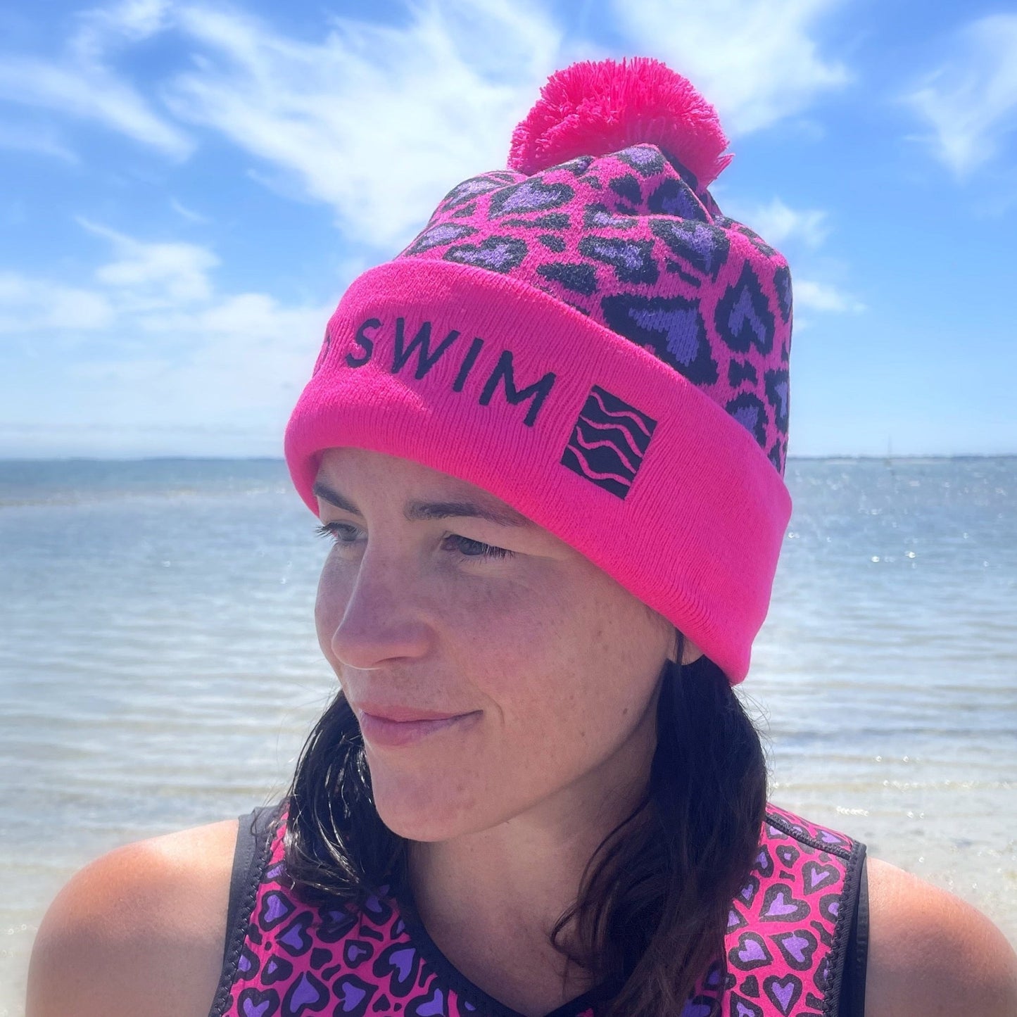 Person wearing a pink leopard print beanie hat with 'Swim' text, looking to the side outdoors by the sea