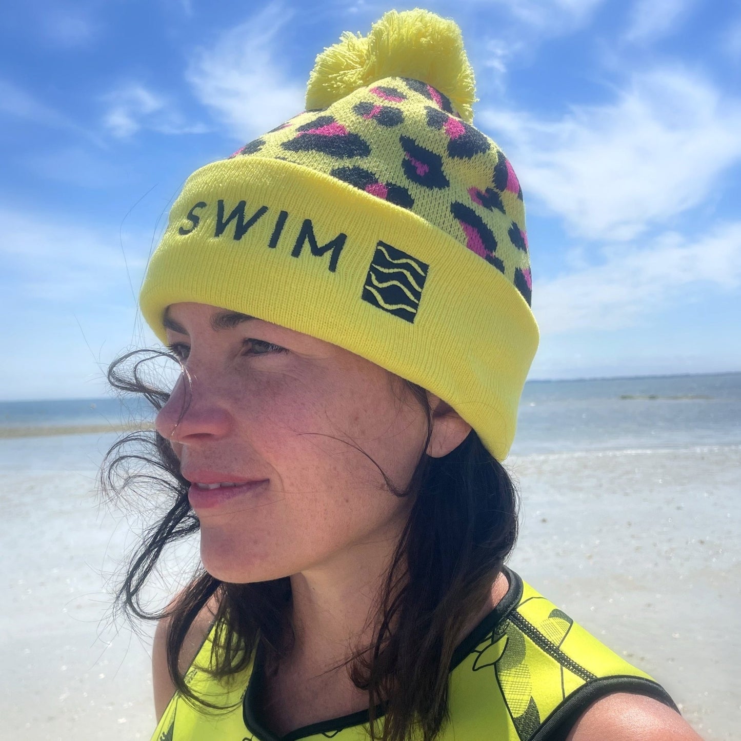 Person wearing a yellow leopard print beanie hat with 'Wild Swim' text, looking to the side on a beach