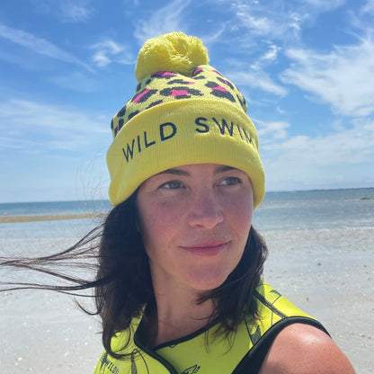 Person wearing yellow leopard print hat and neoprene swimming gear by water.