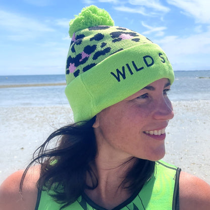 Person wearing a green leopard print beanie hat with 'Wild Swim' text, looking to the side outdoors