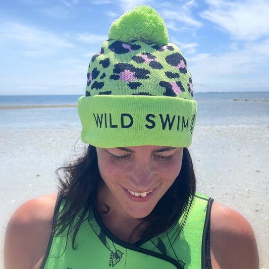 Person wearing a green leopard print beanie hat with 'Wild Swim' text, smiling outdoors by the sea