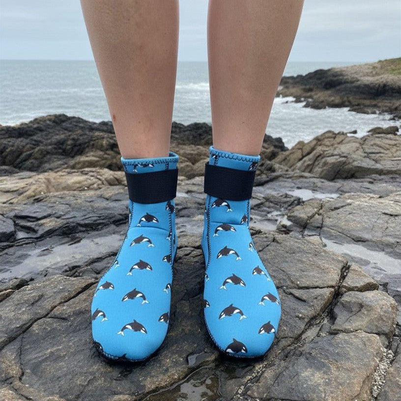 Close-up of blue neoprene swimming socks with black orca print, standing on rocks near the shore