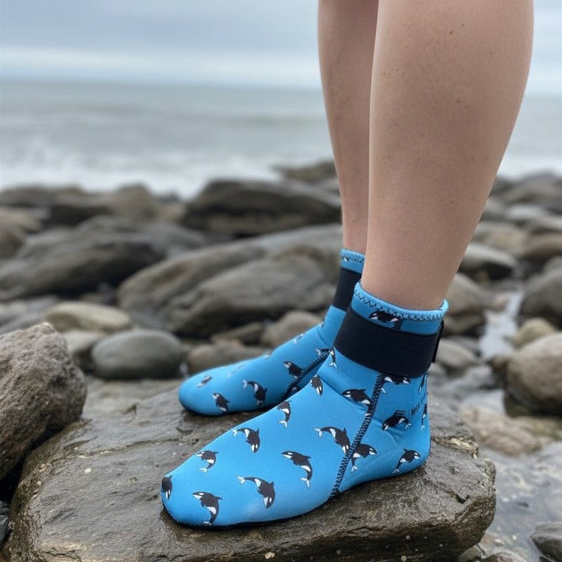 Person wearing blue neoprene swimming socks with black orca print, standing on rocks by the sea